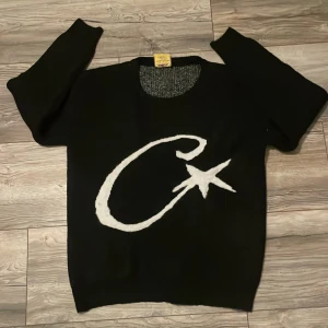 Svart stickad tröja med vit stjärna - Cool svart stickad tröja från PVS i storlek S. Framsidan har en stor vit grafisk design med en stjärna och ett C. Tröjan har rund hals och långa ärmar, perfekt för att ge din outfit en unik touch.
