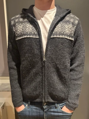 Mörkgrå stickad hoodie från Woolrich - Säljer en mörkgrå stickad hoodie från Woolrich med vit snöflingemönster över axlar, armbågar och huva. Tröjan har dragkedja framtill, huva och långa ärmar. Perfekt för kyliga dagar och har en skön loose passform.