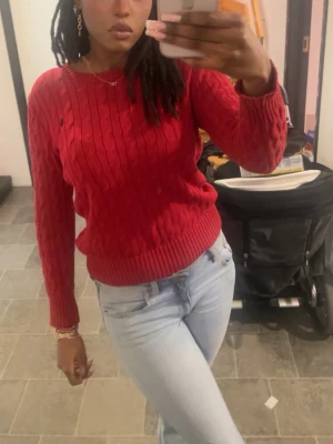 Röd kabelstickad tröja från Ralph Lauren - Säljer en röd kabelstickad tröja från Ralph Lauren med rund halsringning och långa ärmar. Tröjan har klassisk kabelstickad struktur och ribbade muddar vid ärmslut och nederkant. Perfekt för dig som gillar en tidlös och snygg look.