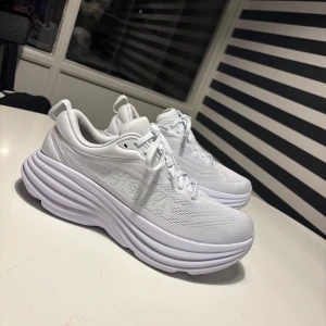 Vita Hoka bondi 8 sneakers strl 40⅔ - Säljer ett par fräscha vita Hoka bondi 8 sneakers med riktigt chunky plattformssula och snyggt mönstrad mesh-överdel. Skorna har snörning och rund tå, perfekt för dig som gillar sportig stil och maxad komfort. Märket syns tydligt på sidan och insidan.