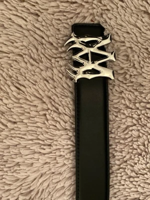 Svart skärp med silverfärgat spänne - Säljer ett svart skärp med ett riktigt coolt och unikt silverfärgat spänne i metall. Skärpet har en stilren design och är tillverkat i slätt material som ger en snygg look. Perfekt för att lyfta din outfit med en edgy detalj.