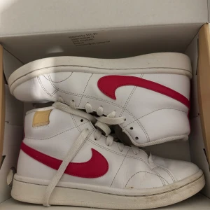 Nike Blazer Mid vit/röd sneakers - Säljer ett par Nike Blazer Mid sneakers i vitt skinn med röd swoosh på sidorna. Klassisk retrostil med högt skaft och snörning. Perfekt för dig som gillar streetwear och vill ha en ikonisk look. Skorna har rund tå och platt sula.