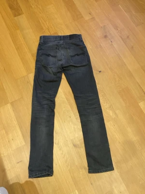 Mörkgrå Grim Tim Nudie Jeans, storlek W27/L30 - Säljer ett par mörkblå jeans från Nudie Jeans i storlek 27/30.snygga detaljer på bakfickorna och en rak passform. Materialet är slitstarkt jeans och färgen har en cool tvättad look med lite skuggningar.