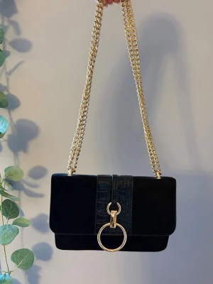 Svart axelväska med guldkedja - Snygg svart axelväska med bred guldkedja och krokodilmönstrad detalj på locket. Väskan har en stor guldfärgad ring framtill. .