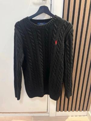  Polo Ralph Lauren tröja - Svart kabelstickad tröja från Polo Ralph Lauren med rund hals och röd broderad logga på bröstet. Tröjan har långärmad passform och ribbade muddar vid ärmslut och nederkant. Perfekt för dig som gillar klassisk stil med en modern touch. Kom dm för eventuella frågor 🧐pris kan diskuteras 💯😊