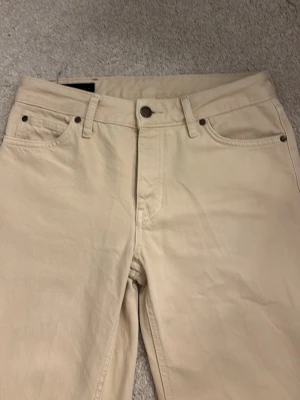 Beige Zara jeans med knappar - Säljer ett par Zara jeans i beige färg. Byxorna har rak passform, fem fickor och knäppning med knappar framtill. Tillverkade i slitstark bomull och har den ikoniska läderpatchen bak i midjan. Perfekt för dig som gillar tidlös och clean stil.