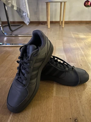 Grå Adidas sneakers  - Säljer ett par svarta Adidas sneakers med klassiska tre ränder på sidan. Skorna har snörning, rund tå och platt sula. Ovandelen är i syntetmaterial och insidan är vadderad för extra komfort. Perfekt för dig som gillar stilrena och sportiga skor.