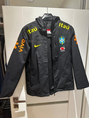 Svart Brasil Nike vindjacka L - Svart vindjacka från Nike med Brasil CBF-logga, gula och orange tryck. Jackan har huva, dragkedja och långa ärmar. Jackan är mindre i storlek. Så om du är 170-177 cm lång passar den dig. Skriv över det minsta lilla!