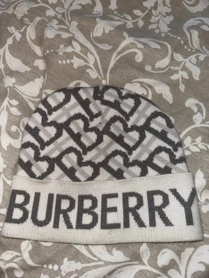 Vit Burberry mössa - Snygg mössa från Burberry i ljusgrått och svart med stort Burberry-tryck framtill. Mössan är stickad i mjukt material. Perfekt för dig som vill ha en trendig accessoar med lyxig känsla.