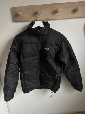 Svart pufferjacka från 8848 Altitude - Säljer en svart pufferjacka från 8848 Altitude med hög krage och dragkedja framtill. Jackan har två fickor med dragkedja och justerbar dragsko nertill. Perfekt för kyliga dagar och riktigt snygg till streetwear-stilen.