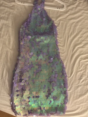 Lila paljettklänning med halterneck - Superfin lila kortklänning med halterneck och stora skimrande paljetter i grönt och lila. Klänningen har en tight passform och är ärmlös, perfekt för dig som vill sticka ut. Materialet är syntet med paljettöverdrag som ger en glittrig effekt. Har bara använts ett år på nyårsafton ☺️är i stl xxs men fassar även xs. Köpte för 700kr