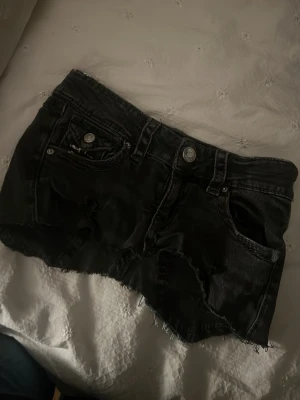 Svart jeanskjol med flaggdetalj - Cool svart jeanskjol med rå kant och slitningar. Framtill har kjolen klassiska fickor och nitar, baktill finns två fickor varav en har Union Jack-tryck och den andra är i blå denim. Perfekt för dig som gillar edgy streetstyle.