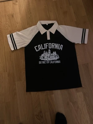 Svart och vit California skjorta - Säljer en svart och vit skjorta med trycket 'California District of California' och en stor vit krage. Skjortan har korta ärmar med svarta ränder och en grafisk illustration framtill. Perfekt för dig som gillar streetwear och college-vibe.