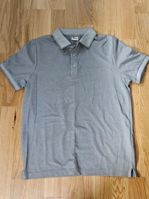 Grå pikéskjorta från AC&Co, S - Oanvänd snygg grå pikéskjorta från AC&Co i slim fit. Skjortan har korta ärmar, klassisk krage med diskreta ränder och tre knappar framtill. Materialet är mjukt och känns som bomull, perfekt för en chill och stilren look.