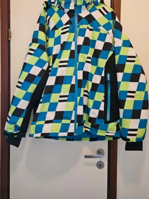 Färgglad rutig snowboardjacka med huva  - Säljer en vindjacka med rutigt mönster i blått, grönt, svart och vitt. Jackan har huva, dragkedja framtill och praktiska fickor med dragkedja. Insidan har snölås med knappar och elastisk kant. Perfekt för dig som gillar färg och mönster.