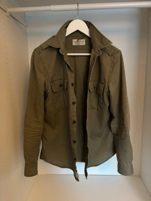 Olivgrön overshirt från Ralph Lauren - Olivgrön overshirt från Denim & Supply Ralph Lauren med klassisk krage, två bröstfickor med knappar och långa ärmar. Jackan är tillverkad i bomull och har en avslappnad passform med knappstängning framtill. Perfekt för lager-på-lager och chill stil.