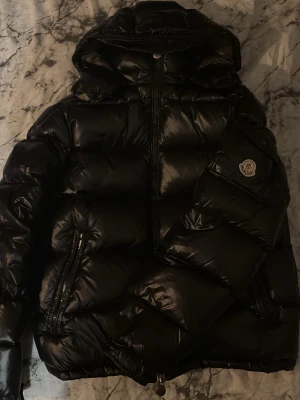 Svart Moncler pufferjacka (ÄKTA) - Säljer en svart pufferjacka från Moncler med glansig finish och huva. Jackan har två snedställda fickor med dragkedja, hög krage och Moncler-logga på ärmen. Perfekt för kalla dagar och har en riktigt fet streetstil.