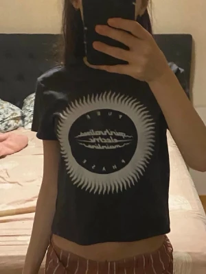 Svart croppad t-shirt med tryck - Svart croppad t-shirt från New Yorker med ett stort vitt grafiskt tryck framtill som ser ut som en sol eller stjärna. Rund halsringning och korta ärmar. Perfekt för dig som gillar streetstyle och vill ha en edgy vibe.