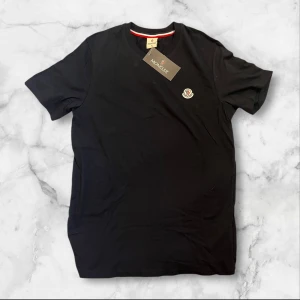 Svart t-shirt från Moncler - Svart t-shirt från Moncler med rund hals och korta ärmar. Diskret Moncler-logga på bröstet och en liten röd detalj på insidan av halsen. Tillverkad i mjuk bomull som känns skön mot huden. Perfekt för dig som gillar stilrena och klassiska plagg.