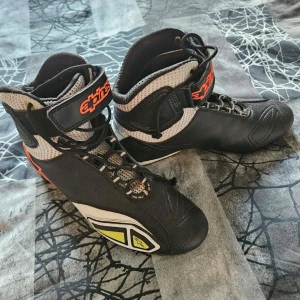 Svarta racingkängor från Alpinestars - Säljer ett par svarta racingkängor från Alpinestars med orange och vita detaljer. Skorna har snörning och kardborreband upptill, samt ventilerande meshpartier på sidorna. Perfekt för motorsport och racing. Snygg design med grafiska inslag och robust konstruktion.
