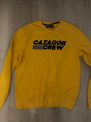 Gul sweatshirt från Cazador Crew - Säljer en gul sweatshirt från Cazador Crew i storlek M. Tröjan har tryckt svart text på bröstet och klassisk rund hals. Materialet är mjuk bomull och modellen är normal passform med långa ärmar och ribbade muddar vid ärmslut och nederkant.