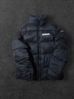 Svart pufferjacka från Napapijri - Svart pufferjacka från Napapijri med stor logga på ryggen och mindre logga på bröstet och ärmen. Jackan har hög krage, dragkedja framtill och quiltad design. , perfekt för kalla dagar. Snygg och stilren look.