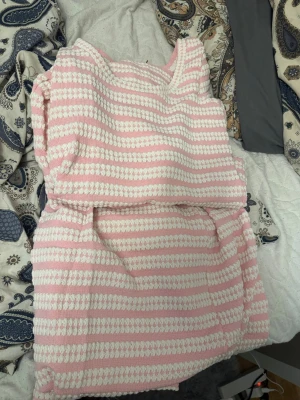 Rosa och vit randig stickad sett Nelly med kjol och tröja  - Supergullig stickad klänning med breda rosa och vita ränder. Klänningen är ärmlös och har en mjuk, mysig känsla med rundad hals. Perfekt för dig som gillar en söt och avslappnad stil. Passar bra till varmare dagar och är lätt att matcha med sneakers eller sandaler.