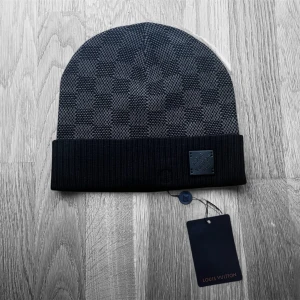 Rutig svart mössa från Louis Vuitton - Snygg svart mössa från Louis Vuitton med klassiskt rutig mönster och ribbad kant. Märket syns tydligt på en läderpatch framtill och på insidan. Materialet är mjukt och stickat, perfekt för kyliga dagar. Mössan har exklusiv känsla och stilren design.
