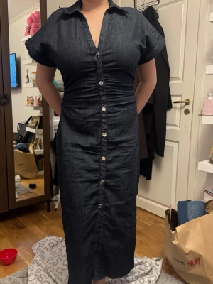 Mörkblå jeansklänning med knappar ZARA - Snygg mörkblå jeansklänning från Zara med v-ringning och korta ärmar. Klänningen har knappar hela vägen framtill och en figurnära passform med rynkade detaljer vid midjan. Perfekt för dig som gillar en stilren och cool look.