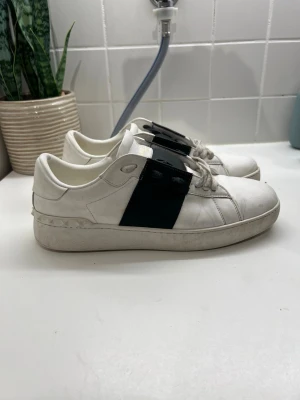 Valentino Garavani vita sneakers - Säljer ett par snygga vita Valentino Garavani sneakers med bred svart läderdetalj över snörningen och coola nitar längs hälen. Skorna har rund tå, platt sula och klassisk snörning. Märkeslogga i guld på plösen. Perfekt för dig som gillar exklusiva sneakers med unik design.