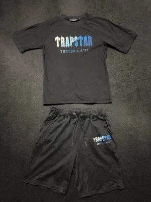 Trapstar svart t-shirt och shorts set - Säljer ett svart set från Trapstar med t-shirt och shorts. T-shirten har ett stort Trapstar-tryck i vit och blå ton på bröstet, och shortsen har matchande logga. Shortsen har snörning i midjan för skön passform. Perfekt för chill eller streetwear.