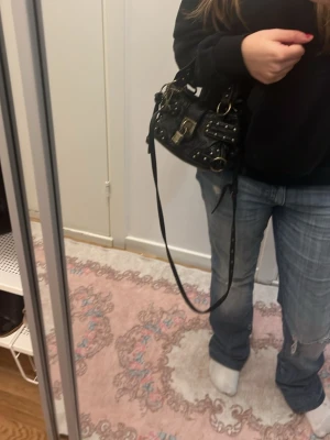 Svart axelväska med metalldetaljer - Säljer en svart axelväska i skinn med coola metalldetaljer och spännen. Väskan har både handtag och lång axelrem, samt dekorativa nitar och en stor låsdetalj framtill. Perfekt för dig som gillar edgy stil och vill ha plats för det viktigaste.