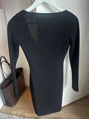 Svart glittrig klänning XS från Hm - Snygg svart glittrig klänning från Hm i storlek XS. Klänningen har lång ärm och en djup rundad urringning bak. Materialet är stretchigt och glittrar diskret i ljuset, vilket ger en cool och trendig vibe. Perfekt för dig som gillar att sticka ut med stil.