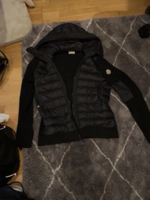 Svart hoodie från Moncler med huva - Svart hoodie från Moncler med quiltad front och huva. Tröjan har långärmade stickade partier och dragkedja framtill. Moncler-logga på ärmen och praktiska fickor med dragkedja. Perfekt för kyliga dagar och snygg till streetwear.