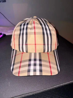 Burberry rutig keps i beige och svart - Säljer en snygg Burberry keps med klassiskt rutigt mönster i beige, svart, vitt och röda detaljer. Kepsen har justerbar snapback och Burberry London England-etikett baktill. Materialet är bomull och polyester, vilket gör den både bekväm och stilren. Pris går att diskutera