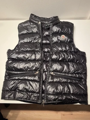 Moncler Gui Gilet Väst - En väst som är välanvänd, finns ett litet hål, se bild 2.           Sätt en svart liten tejpbit över det så är det inget som syns på håll. Dragkedjan funkar inte, därav säljs denna billigt till någon som kanske är på en tajt budget🙂