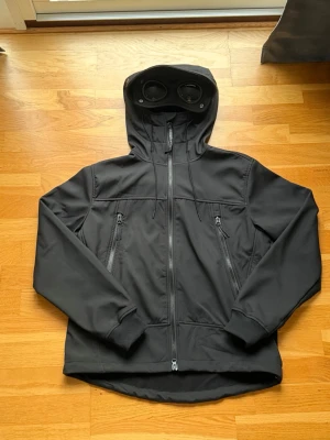 Svart vindjacka från C.P. Company - Svart vindjacka från C.P. Company med ikoniska goggles i huvan. Jackan har två snedställda fickor med dragkedja, ribbade muddar och hel dragkedja framtill. Materialet är syntet och jackan har en clean, modern look som sticker ut med detaljerna på huvan.