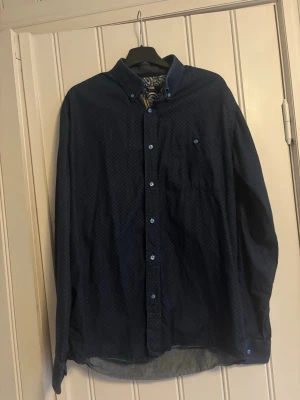 Mörkblå skjorta från STCRM - Snygg mörkblå skjorta från STCRM med diskret mönster och paisley-mönstrad insida vid kragen. Skjortan har klassisk krage, bröstficka och knappar framtill. Perfekt för dig som gillar detaljer och vill ha en stilren look.