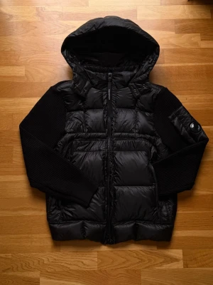 Svart pufferjacka från C.P. Company - Svart pufferjacka från C.P. Company med huva och ribbstickade ärmar. Jackan har en snygg quiltad look, dragkedja framtill med logotyp och en ikonisk linsdetalj på ärmen. Materialmix av polyester och stickad textil gör den både varm och stilren.