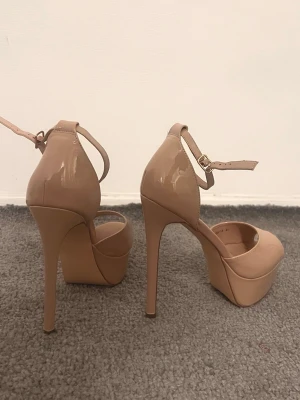 Beige platåpumps med hög klack - Säljer ett par beige pumps med platåsula och riktigt hög klack. Skorna har rund tå, öppen tå och ett smalt ankelband med spänne. Ytan är glansig och ger en snygg finish som sticker ut. Perfekta för dig som gillar att ta ut svängarna med höjden. Oanvända.