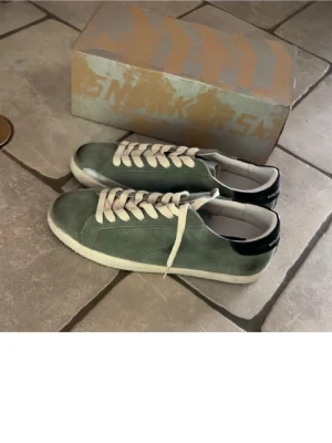 Gröna Golden Goose - Säljer ett par snygga gröna sneakers från Golden Goose med vit sula och vita snören. Skorna har en rund tå och är tillverkade i mocka med svart detalj på hälen. Perfekta för dig som gillar stilrena och coola sneakers. Dem är lite slitna under till. Men annars riktigt bra skick. Nypris ca 4800kr mitt pris 1149. Priset är ej hugget i sten. Hör gärna av er vid minsta lilla fundering. 