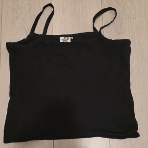 Svart linnetopp från R - En enkel svart topp från R med smala axelband och rak halsringning. Toppen är ärmlös och har en klassisk, stilren design som passar till många olika outfits. Perfekt för varma dagar eller som lager under andra plagg.
