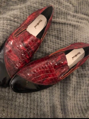 Vinröda loafers från Topman i skinn - Säljer ett par riktigt snygga vinröda loafers från Topman i storlek 42. Skorna har ett glansigt krokodilpräglat mönster, svarta lackdetaljer på tån och orangea sömmar. Tillverkade i skinn och har en klassisk, elegant form med låg klack.