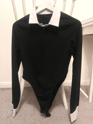 Svart byxdress med vit krage - Snygg svart byxdress med lång ärm, vit krage och vita muddar vid ärmsluten. Kroppsnära passform och dragkedja framtill. Perfekt för dig som gillar stilrena kontraster och vill sticka ut med en cool look.