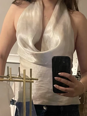 Champagnefärgad omlott halterneck topp - Säljer en champagnefärgad halterneck topp med omlott-drapering framtill. Toppen har en glansig finish och är ärmlös, perfekt för att sticka ut. Materialet känns lyxigt och har en lätt skimrande yta som ger en elegant vibe.