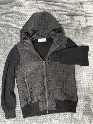 Svart hoodiejacka/cardigan från Dior - (Pris går att diskutera) Svart hoodiejacka från Dior med quiltad framsida och huva i glansigt material med Dior-mönster. Ärmar och rygg är stickade och ger en schysst kontrast. Jackan har två dragkedjefickor och ribbade muddar. Perfekt för dig som vill ha en unik och lyxig streetstil.