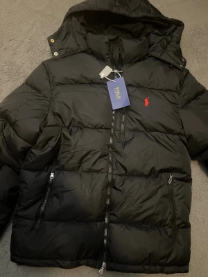 Svart pufferjacka från Polo Ralph Lauren - Svart pufferjacka från Polo Ralph Lauren med röd broderad logga på bröstet. Jackan har huva, flera dragkedjor och en clean, quiltad design. Perfekt för kalla dagar och har en modern, oversized vibe. Materialet är syntet och dun, vilket gör den både lätt och varm.