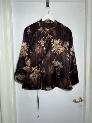 Silkesjacka - Sofistikerad jacka i 100 % siden med ett mjukt, poetiskt blommönster i dämpade mörka toner. Tyget är lätt, följsamt och har en naturlig lyster som ger plagget ett exklusivt uttryck.  Den lätt figurnära men bekväma passformen tillsammans med knytbandet framtill gör att jackan kan justeras efter kroppen. De diskreta dekorativa detaljerna ger en subtil orientalisk känsla utan att bli för markerad.  🧵 Material 	•	100 % siden  📐 Passform 	•	Bekväm modell med justerbart knytband 	•	Lätt att bära både 