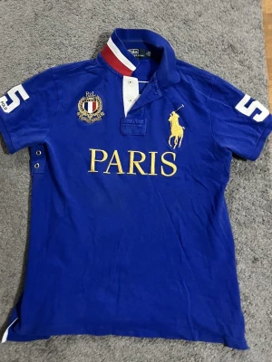 Blå polotopp från Polo Ralph Lauren - Säljer en blå polotopp från Polo Ralph Lauren med gul PARIS-tryck och broderad logga på bröstet. Toppens krage har vita och röda detaljer, och siffran 5 är tryckt på både ärm och rygg. Klassisk kortärmad design i mjuk bomull.