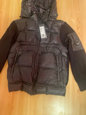 Svart dunjacka från C.P. Company - Säljer en svart dunjacka från C.P. Company med huva och stickade ärmar. Jackan har quiltad front, dragkedja och en cool detalj med logga på ärmen. Perfekt för dig som vill ha en snygg och varm jacka med streetkänsla. Storlek M passar S